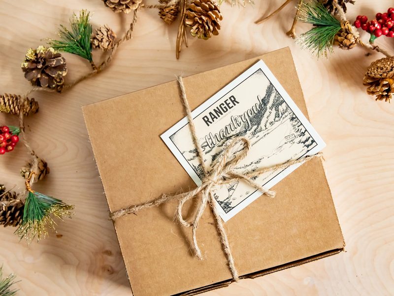 A kraft gift box tied with twine and a Ranger label among pinecones and holiday greenery